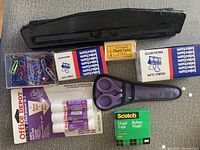 Photo showing a tray with a variety of office supplies including small colored paper clips, two boxes of binder clips, a box of thumb tacks, purple scissors, two glue sticks, and a roll of Scotch Magic Tape.