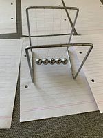 Metal Newton's Cradle device with five pendulum balls hanging from a metal frame, placed on lined paper.