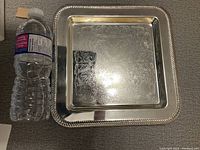 Top view of rectangular silver plated tray showing ornate engraved floral pattern and beaded edge, placed next to water bottle for size reference.