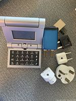 Photo showing a portable electronic device with a numeric keypad and small screen alongside four different travel power adapters/plugs for various plug types.