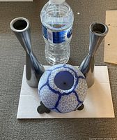 Two silver-toned metal candle holders with tapered tops standing upright beside a blue-and-white round ceramic candle depicting a crackle pattern, with a water bottle for scale.