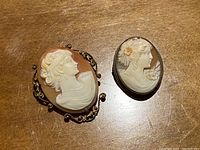 Two cameo pins shown side by side on a wood surface, both featuring carved white female profiles on brown background.