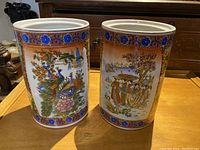 Two cylindrical Chinese vases with colorful painted designs, one showing a peacock on a branch and the other a group of figures, sitting side by side on a wooden table.