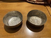 Two silver bowls on a wooden surface, showing engraved floral and vine patterns inside and outside.