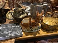 Photo showing the front view of assorted ceramic, metal, and decorative household items on a wooden table.