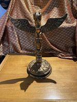 Front view of bronze totem sculpture showing carved vertical totem with broad wings at top and round base.