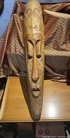 Front view of elongated wooden African mask with carved eyes, nose, lips, and surface patterns, placed on a wooden table with patterned fabric background.