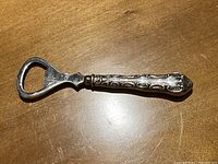 Full view of the silver bottle opener showing the metal open-end and ornate handle against a wood surface.