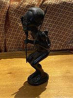 Front-left angle view of the carved wooden African statue showing the facial features, crouched posture, and hand holding a staff or spear.