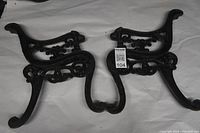 Both black cast iron bench ends shown side by side on white fabric background.