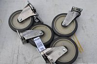 Four heavy duty casters laid out, metal mounting plates, grey rubber wheels, one locking caster visible.