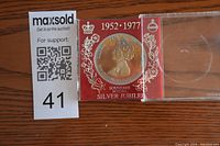 Clear plastic case containing the Queen Elizabeth II Silver Jubilee medal against a red background, showing the front profile of the Queen with decorative elements.