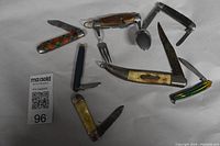 Overview of 7 vintage pocket cutting tools laid out on white background showing various blade sizes and handles, including small child's utensil pocket knife with fork and spoon.