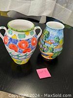Photo showing both vases side by side on black table, colorful floral/geometric large vase with handles on left, village scene smaller vase on right