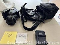 Camera, battery charger, camera case and neck strap displayed on a white surface, showing the camera's front and lens.