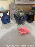 Four vases displayed on white surface including Murano, rustic two-handle, black ceramic-metal, and white ceramic vases.