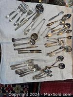 Image showing arranged assorted silver plated forks, spoons, knives, tongs, and small ladle with mother of pearl handle laid out on white cloth.