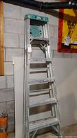 Front view of 6 ft aluminum step ladder leaning against cinder block wall, showing paint splatters on steps and frame.