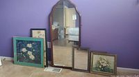 Overall view of three framed prints, needlepoint artwork, and two mirrors leaning against purple wall and floor.