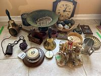 Overall view of all items on floor including basket with marble eggs, metal bowl, multiple bells, figurine, clock, thermometers, barometer, wooden box, tankard.