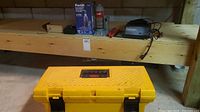 Wide angle showing the yellow plastic toolbox, Pro-Lift hydraulic bottle jack with original box, and Eliminator charger side by side on a wooden shelf in a basement.
