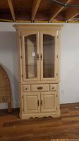 Front view of the light wood corner cabinet showing the glass doors, drawer, and lower doors.