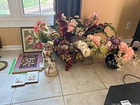 Wide shot showing multiple faux floral arrangements set on floor, framed art leaning against wall including pressed flower plaques and fall leaf print, and a musical unicorn figurine on the floor.