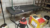 Front angled view of black plastic folding table showing foldable metal legs and surface condition, with vacuum and other items beneath.