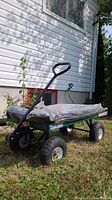 Full side view of green metal garden wagon loaded with a bag of black mulch, positioned on grass by white house wall with window above.