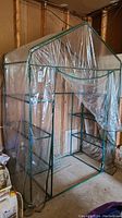 Greenhouse with clear plastic cover and green metal frame, showing front and side with shelving and zipper door.