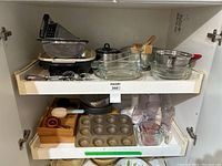 View of open kitchen cabinet with two shelves containing various kitchenware items including metal strainers, graters, glass mixing bowls, baking pans, and other utensils.