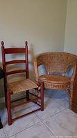 Two chairs: red wooden ladder back chair with woven cane seat, cane tub chair side by side on tiled floor.