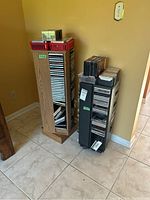 Two media storage stands in corner, one wooden swivel stand with CDs and cassette tapes, smaller black stand on casters with CDs and VHS tapes.