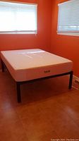 View of Lucid mattress on black metal bed frame from longer side in orange room with tile floor and white window blinds.