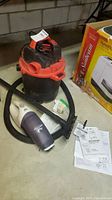 Craftsman shop vac with red and black body, hose and extension tube, Black & Decker handheld dust buster vacuum, instruction manuals on the floor beside items.