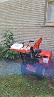 Front view of orange Simplicity snow blower parked against brick wall