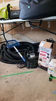 Photo of sump pumps on floor including Red Lion pump box, black hose, pool net and container of chlorine tablets