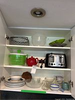 Photo of kitchen cabinet shelves containing all items. Top shelf has metal storage basket and plastic bowl. Middle shelf has salad spinner, red teapot, toaster, and black container. Bottom shelf has stacked plates, bowls, glass bowls, cruet, and assorted serving items.