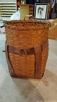 Side view of vintage woven fishing basket showing leather belt with metal buckle and handle on top.