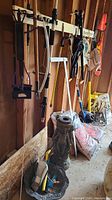 Photo of garden tools including rakes, hoes, brushes, hanging on a wall next to bags of mulch and resin birdbath.