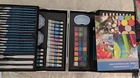 Top-down view of the open artist kit briefcase showing organized paint tubes in oil, watercolor, acrylic, brushes, palettes, pencils, and booklet.