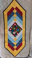 Full view of quilted table runner with geometric colorful pattern and pointed ends in various colors including yellow, red, blue, and black.