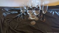 Nine clear glasses arranged on dark fabric, showing three wine glasses with round bowls and six taller champagne flutes with narrow bowls, all with black stems.