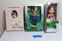 Photo of three dolls in their boxes: My Fair Lady Barbie, Birds of Beauty Peacock Barbie, and Disney Ariel doll with damaged box.