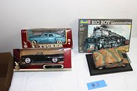Four collectible models including a boxed 1960 Chrysler 300F in blue, black 1948 Tucker car in original packaging, boxed Revell Big Boy Locomotive model kit, and camouflaged Maisto Humvee on display base.
