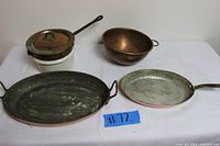 Four cookware items on white surface, including ceramic double boiler with copper lid, copper mixing bowl with brass ring, oval copper fish pan, and large copper skillet
