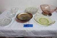 Seven glassware items displayed on a white cloth, including clear, pink, amber, and amethyst glass bowls and plates arranged in two rows