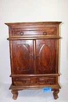 Front view of wooden cabinet showing two carved doors, top decorative drawer, and bottom two smaller drawers