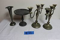 Photo showing two vintage silverplate three-arm candelabras, one silverplate single bud base with flowered top, and two rustic metal candle bases on a white surface.
