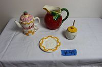 Front view of all five items on white cloth: strawberry pitcher, honey dispenser, tea pot, tea cup, and saucer.